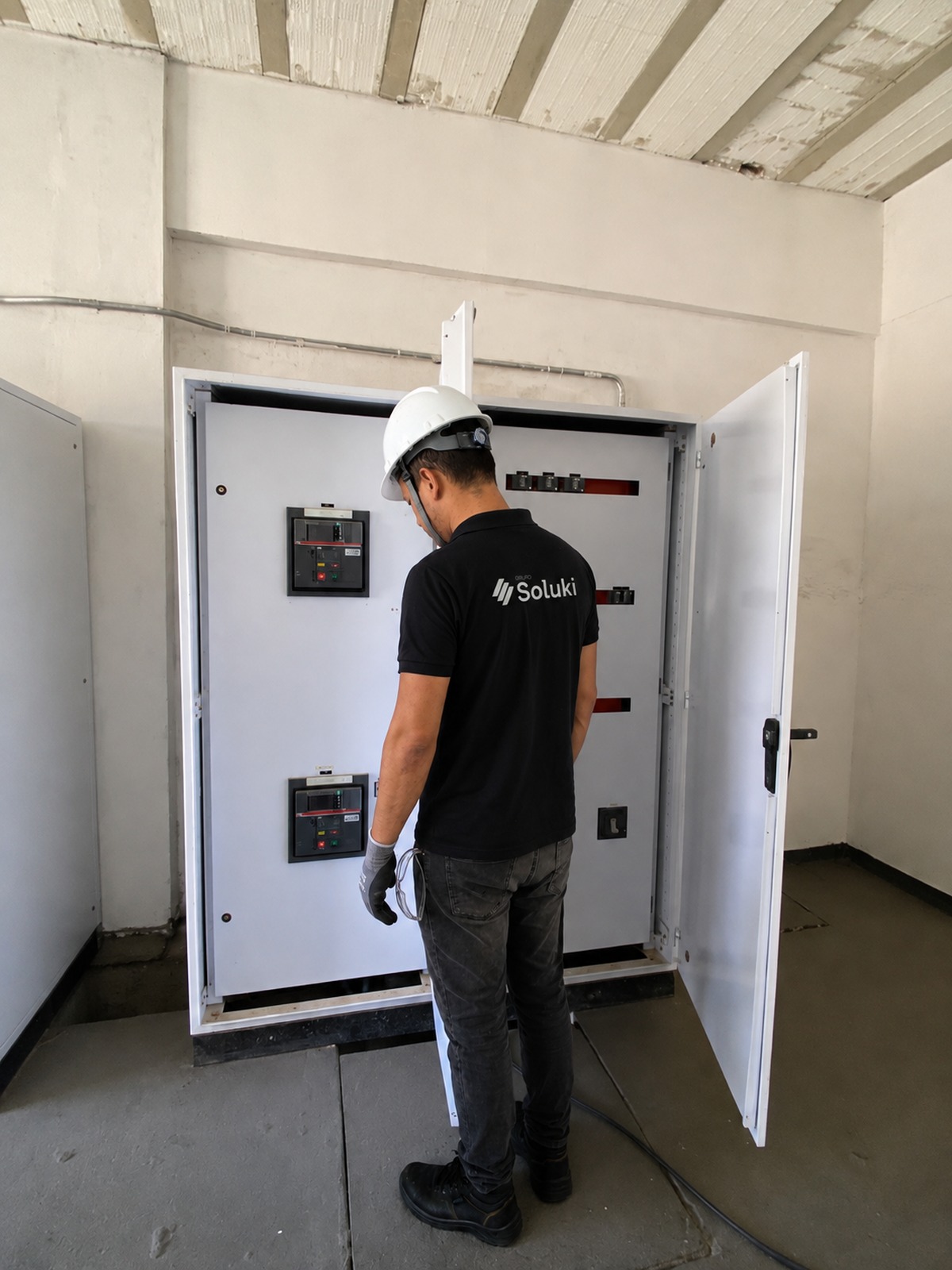 Engenheiro da Soluki, de costas, inspecionando um quadro elétrico com capacete e EPIs